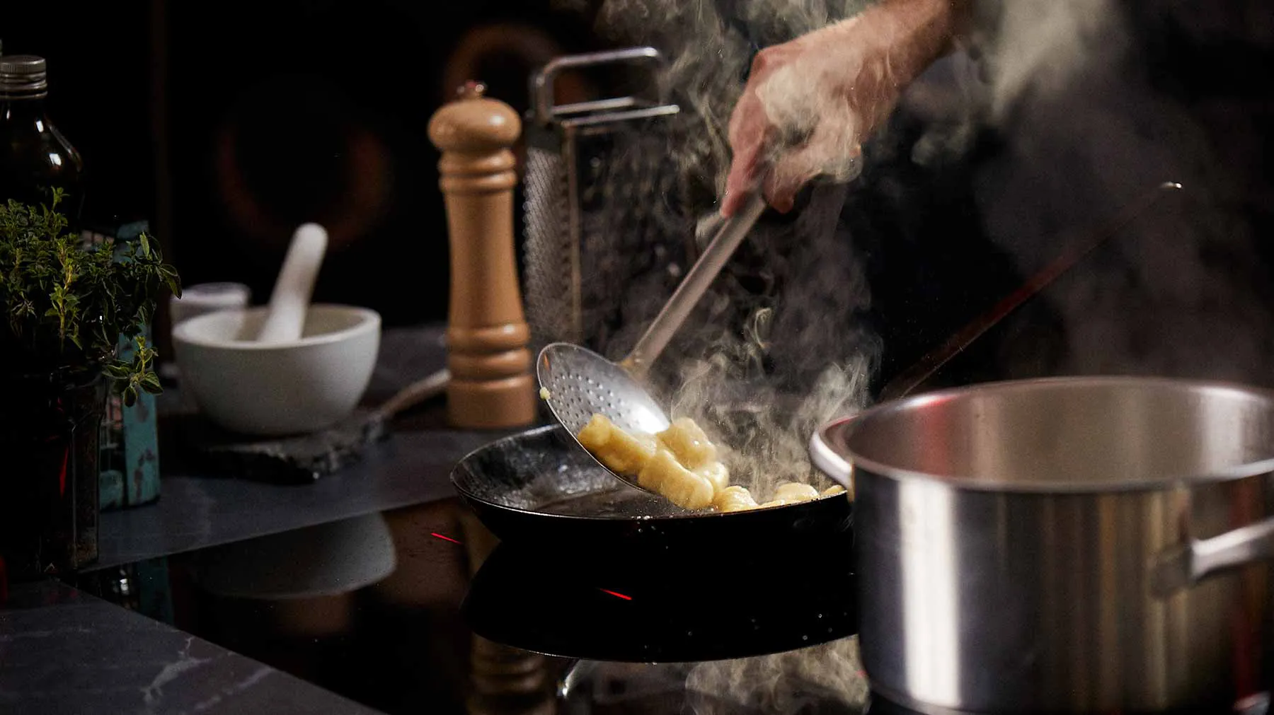 Cooking scene with steaming pan, herbs, and cooking utensils on a dark background.