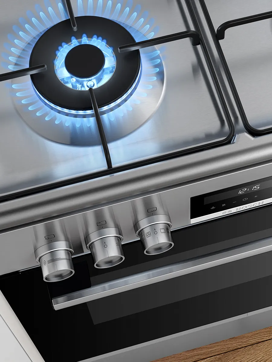 Close-up of a freestanding stainless-steel gas range: a lit burner with blue flame on the cooktop, three metal control knobs and a digital display reading "12:15," and a black oven door with a stainless handle and wooden floor visible at the bottom.