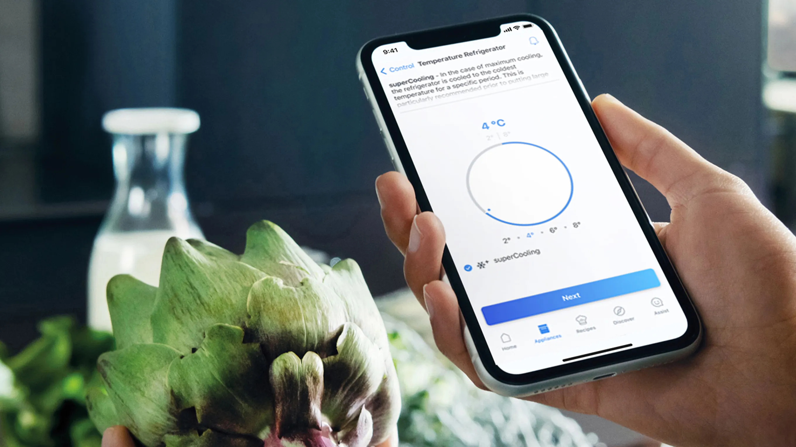 A person uses the Siemens Home Connect app on their smartphone to activate the "superCooling" feature on their smart refrigerator. In the foreground, a fresh artichoke is held, while a bottle of milk sits on the counter in the background.