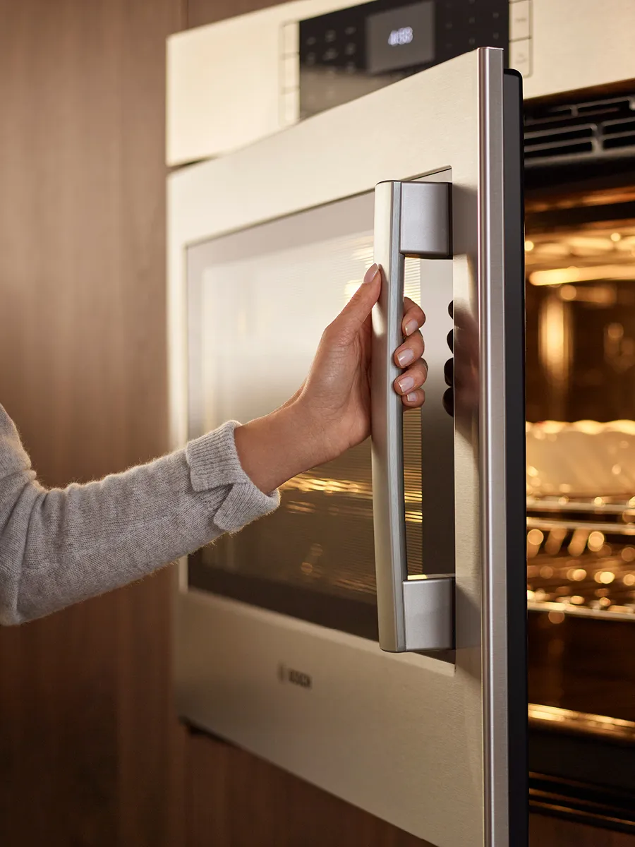 A person in a gray sweater gently pulls the handle of a sleek, modern oven within a contemporary kitchen.