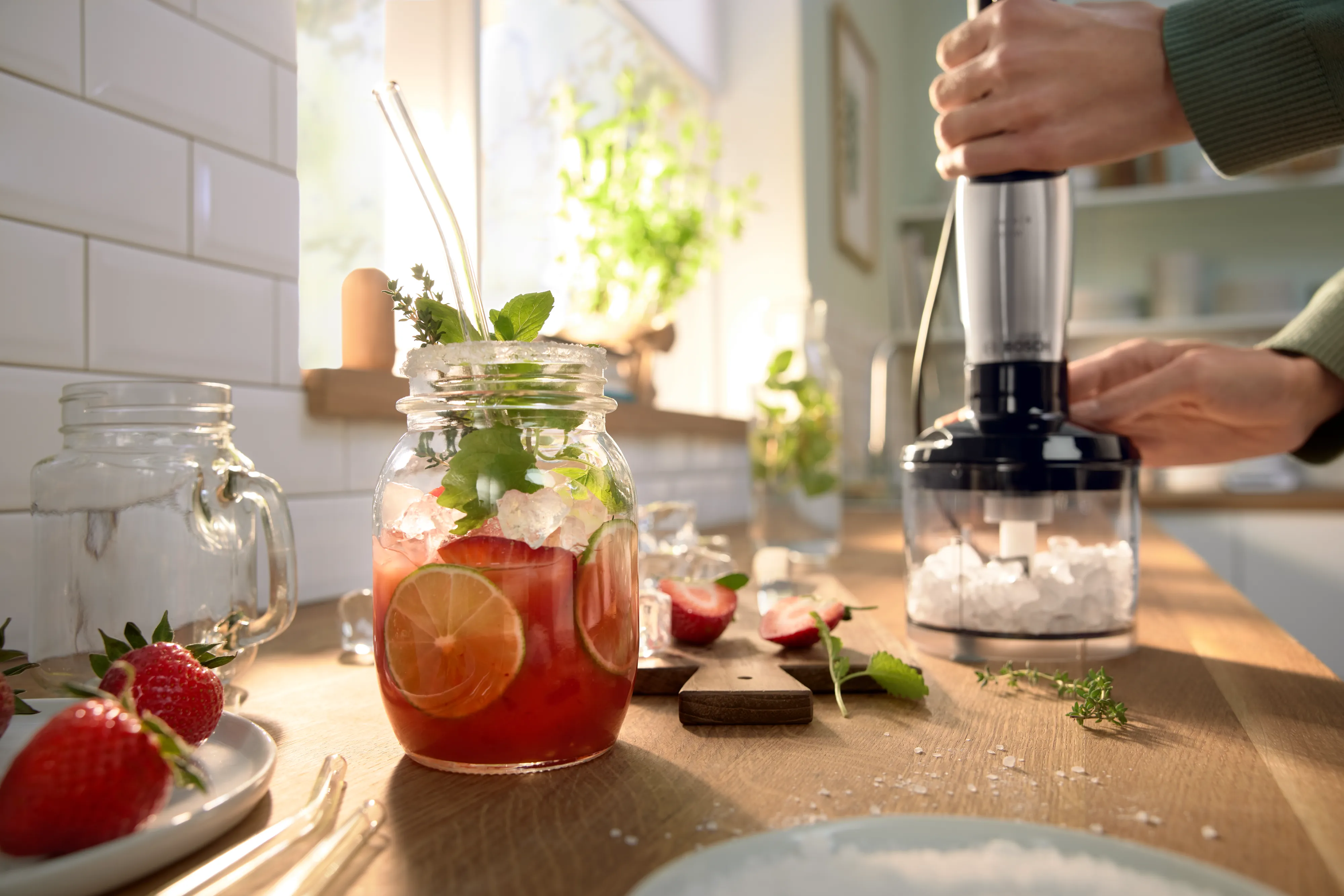 Hands using an immersion blender to crush ice on a kitchen counter, with a jar of iced fruit drink and strawberries nearby.
