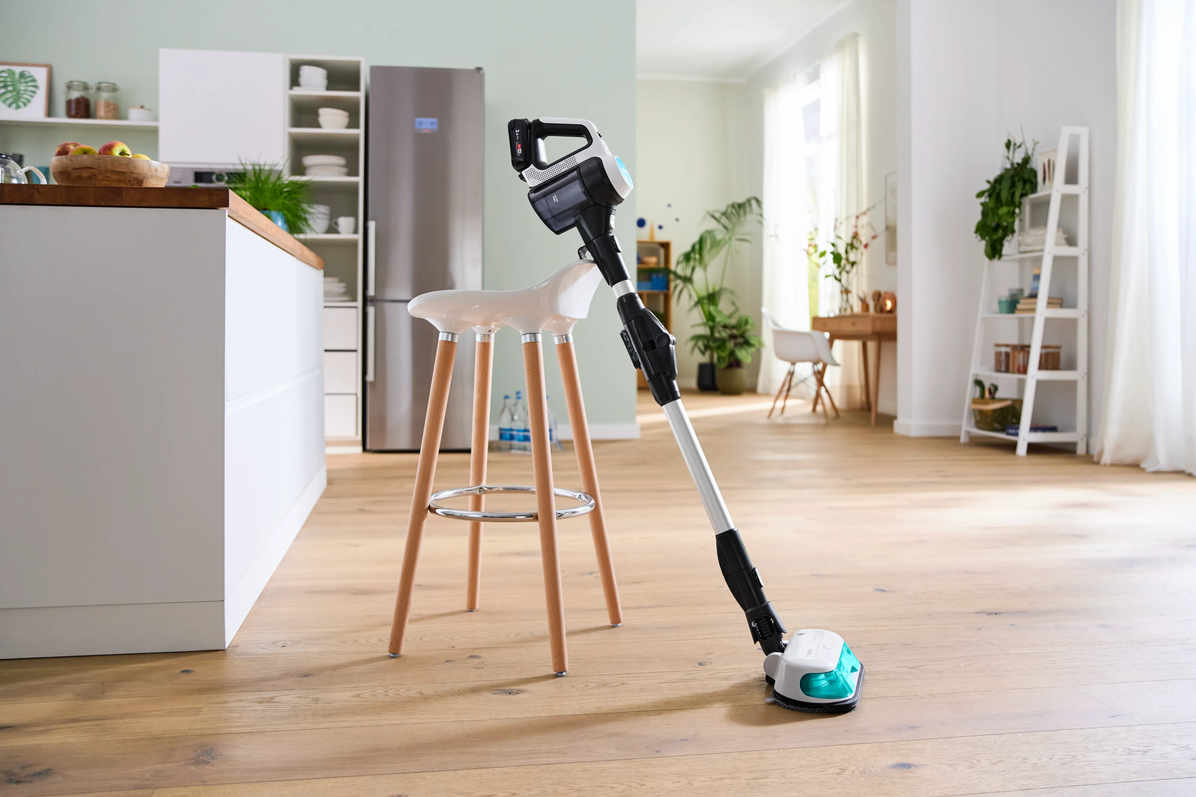 A Bosch cordless wet and dry vacuum cleaner stands upright next to a white and wooden bar stool in a bright, modern kitchen with a refrigerator and plants in the background.