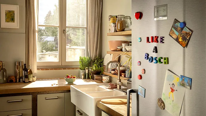 Cuisine chaleureuse avec une fenêtre, des plantes sur le plan de travail et un réfrigérateur décoré de magnets en forme de lettres colorées et de cartes postales.