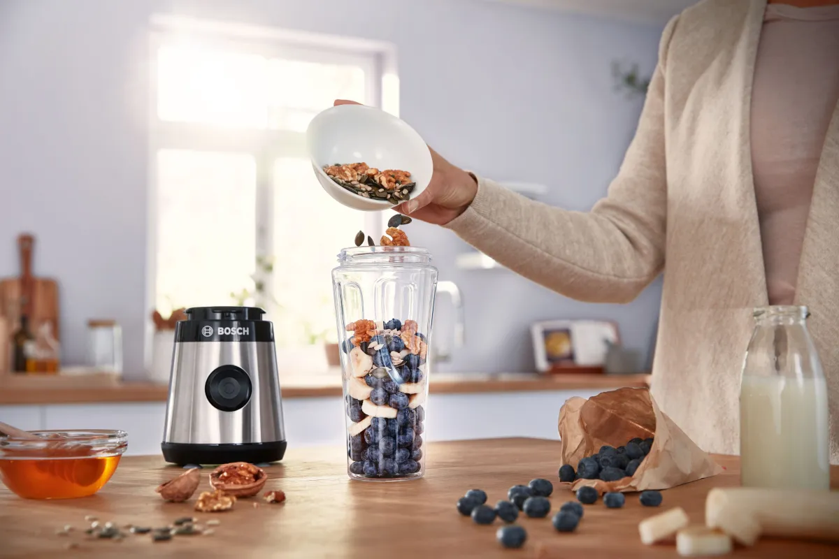 Una persona aggiunge noci e semi a un frullatore pieno di mirtilli e frutta affettata su un piano di lavoro in cucina, con miele e latte nei dintorni.