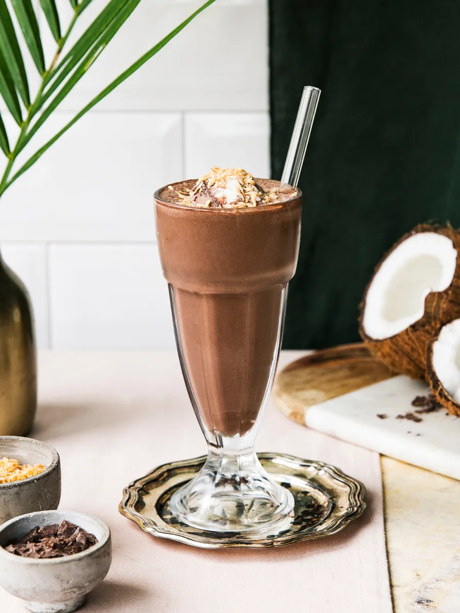 Milkshake de ciocolata cu frisca, nuca de cocos, si batai de ciocolata pe o masa de lemn cu frunze tropicale.