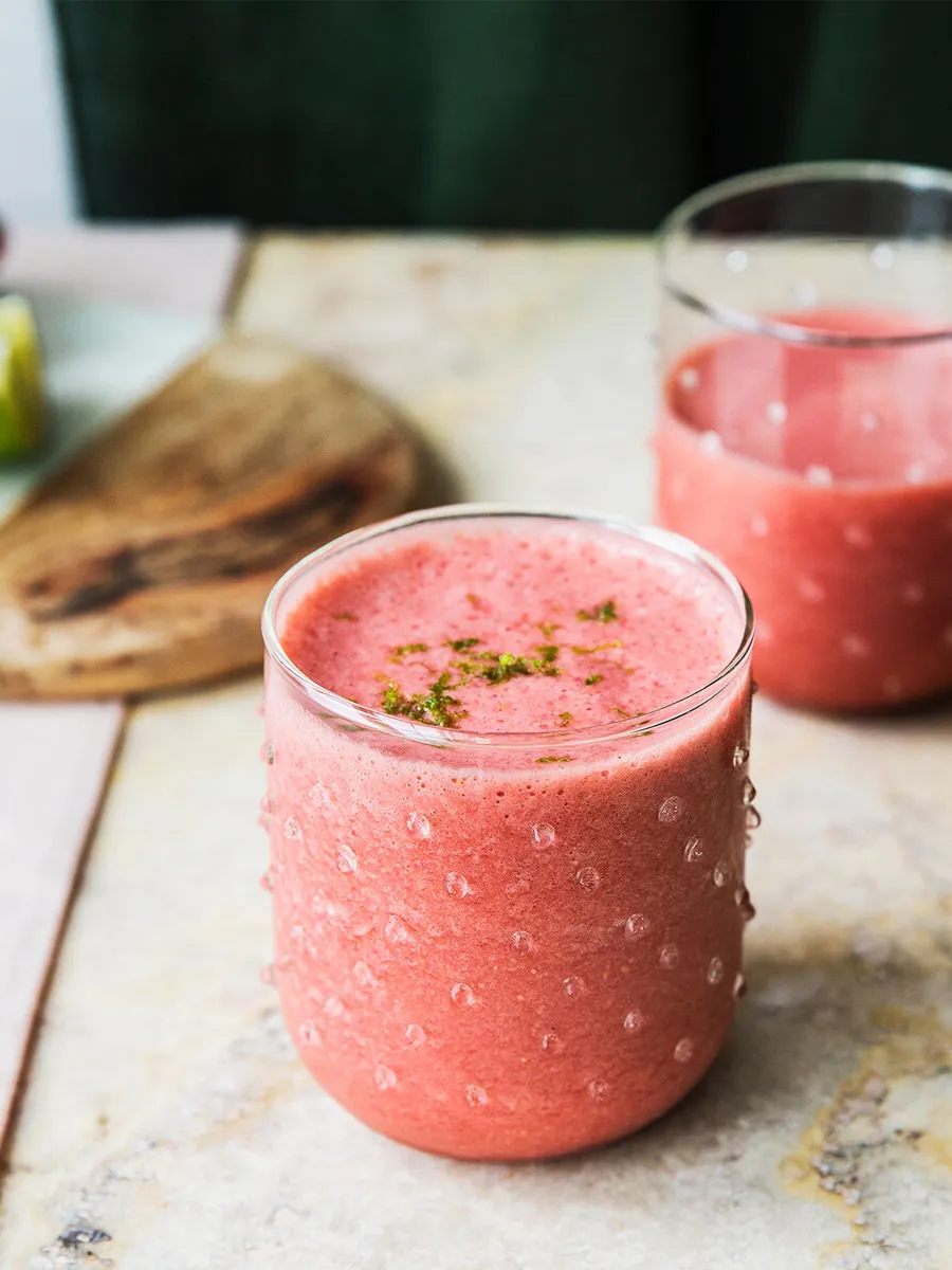 Două pahare cu textură, conținând un smoothie roz, așezate pe o suprafață deschisă la culoare, cu o tăviță din lemn în fundal