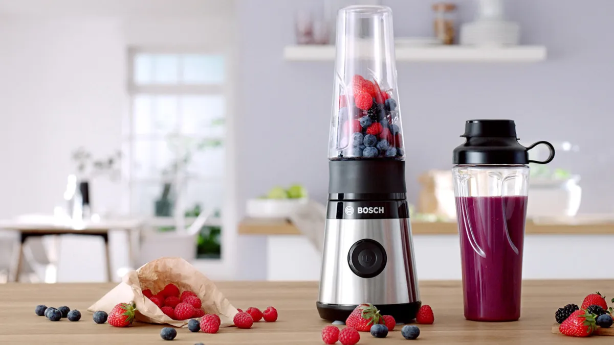 Blender cu fructe de padure proaspete, cupa de piure, și bucătărie interior în fundal.
