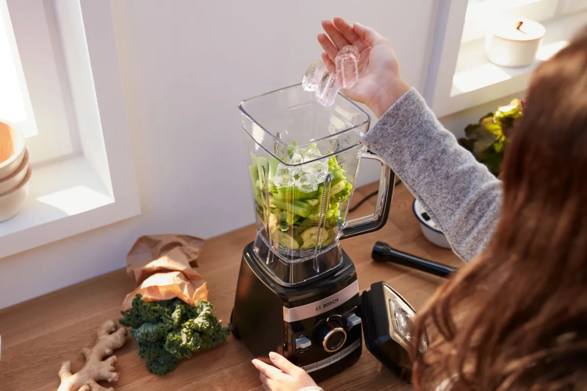 O persoană care pune cuburi de gheață într-un blender umplut cu legume cu frunze verzi, așezat pe blatul din bucătărie