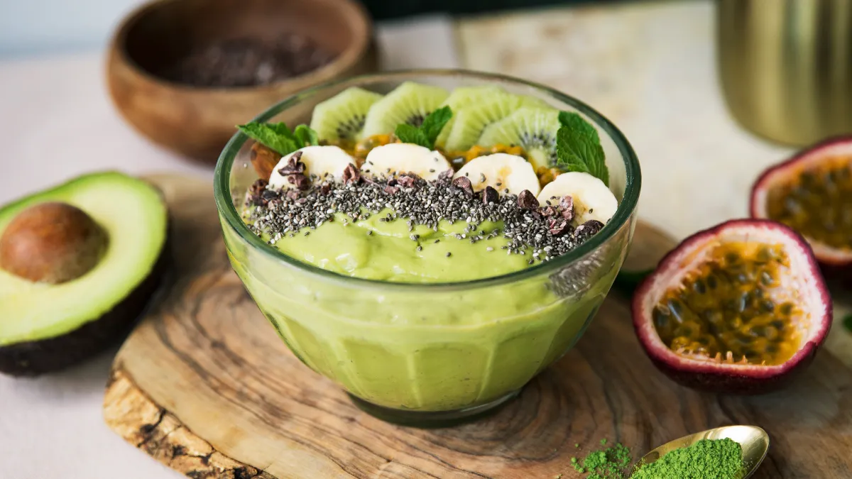 Bol de sticlă cu un smoothie verde, garnisit cu felii de banană, felii de kiwi și semințe de chia, așezat pe o tăviță de lemn și înconjurat de jumătăți de avocado și de fructul pasiunii
