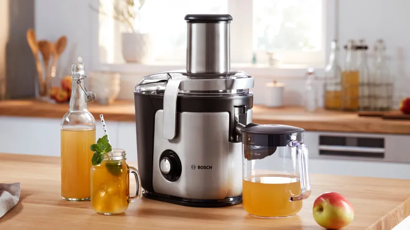 Un extracteur de jus Bosch est posé sur un plan de travail en bois de cuisine, entouré de boissons, d'une pomme fraîche et de bouteilles en verre.