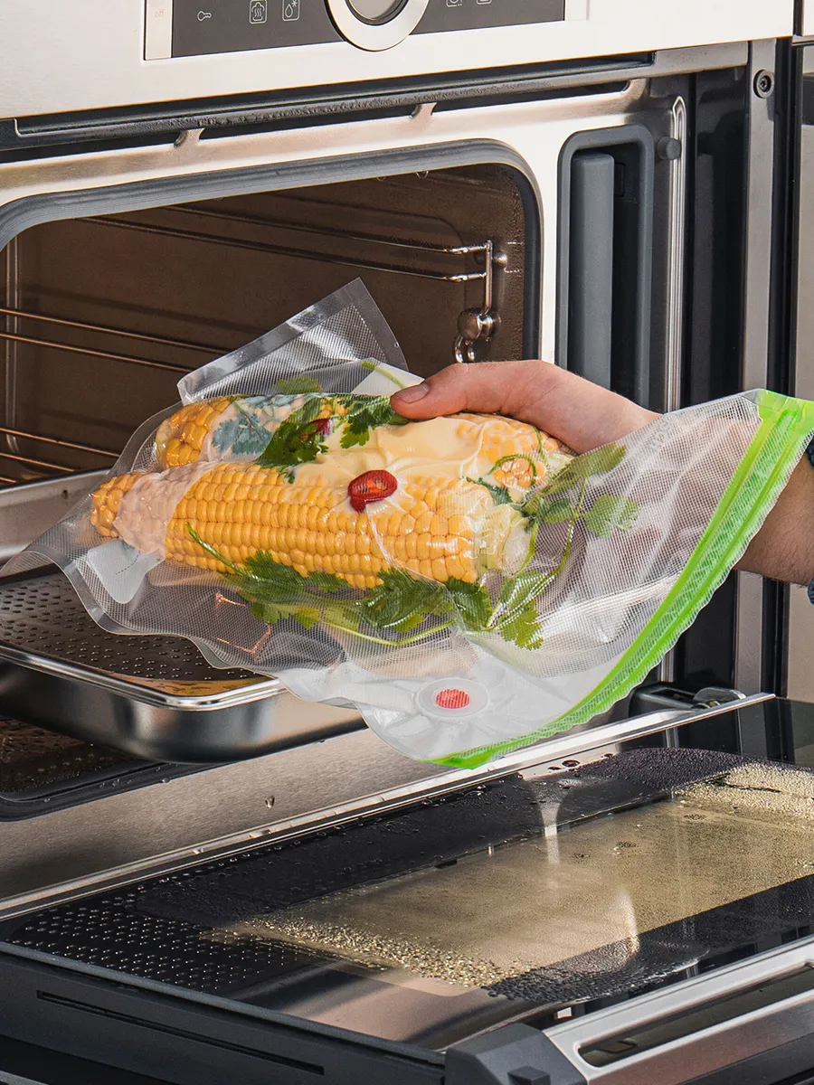 A hand holding a vacuum-sealed bag of corn and herbs is approaching an oven, ready for cooking.