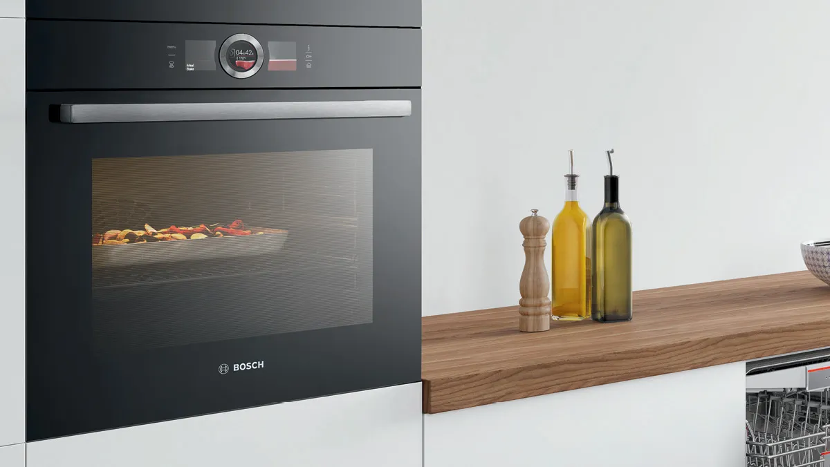 A Bosch oven with a view of colorful vegetables roasting inside, alongside an elegant wooden countertop featuring oil bottles and a pepper mill.