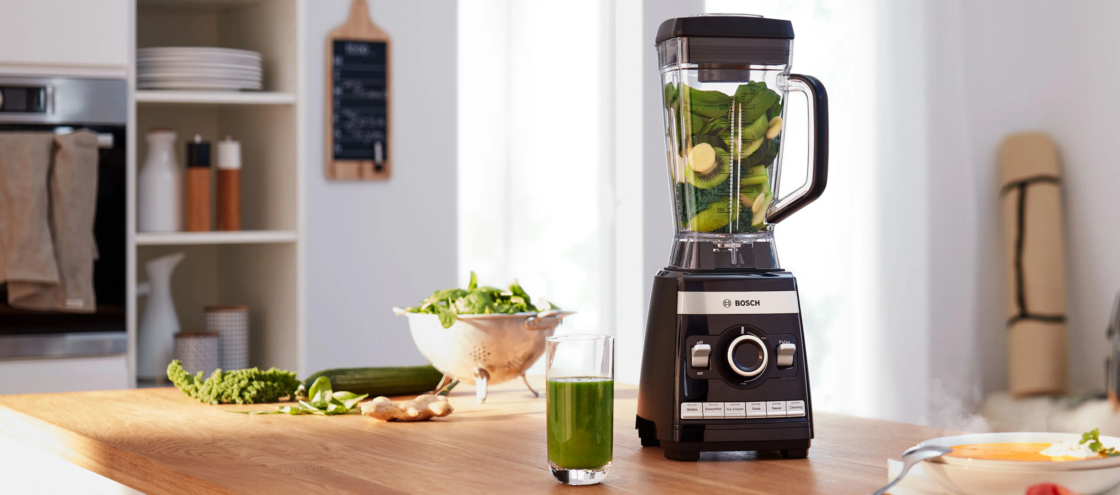 Blender de bucătărie cu recipient transparent, plin cu ingrediente verzi, așezat lângă un pahar cu smoothie verde, pe un blat de bucătărie din lemn