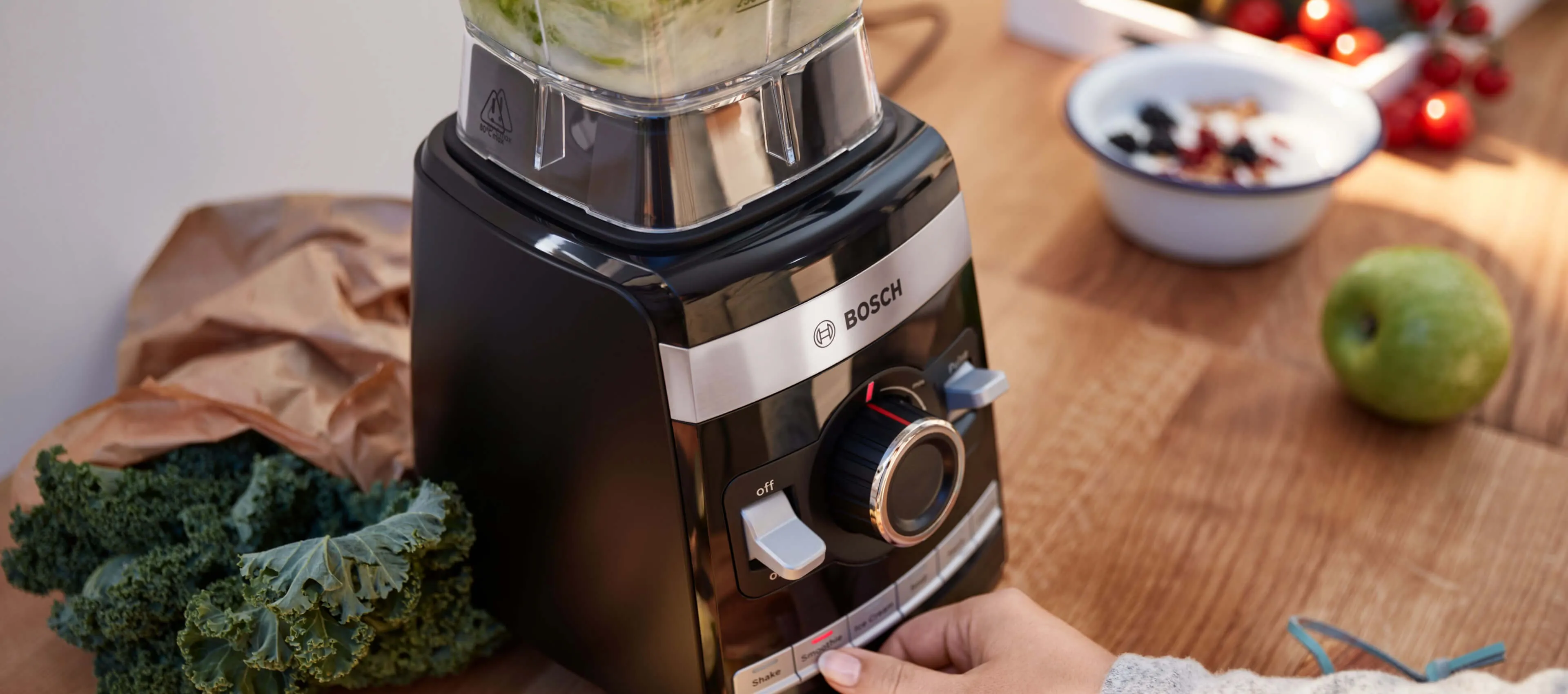 Prim-plan al unui blender negru Bosch VitaBoost Seria 6 așezat pe o masă din lemn, cu varză crețată, o pară și un bol cu fructe de pădure lângă el