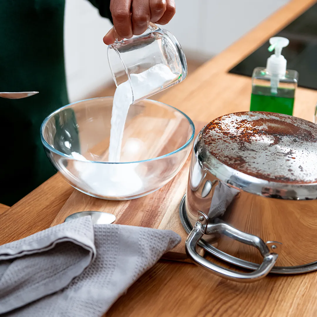 A person pours baking soda and dish soup into a glass bowl beside a stainless steel pot on a wooden counter.
