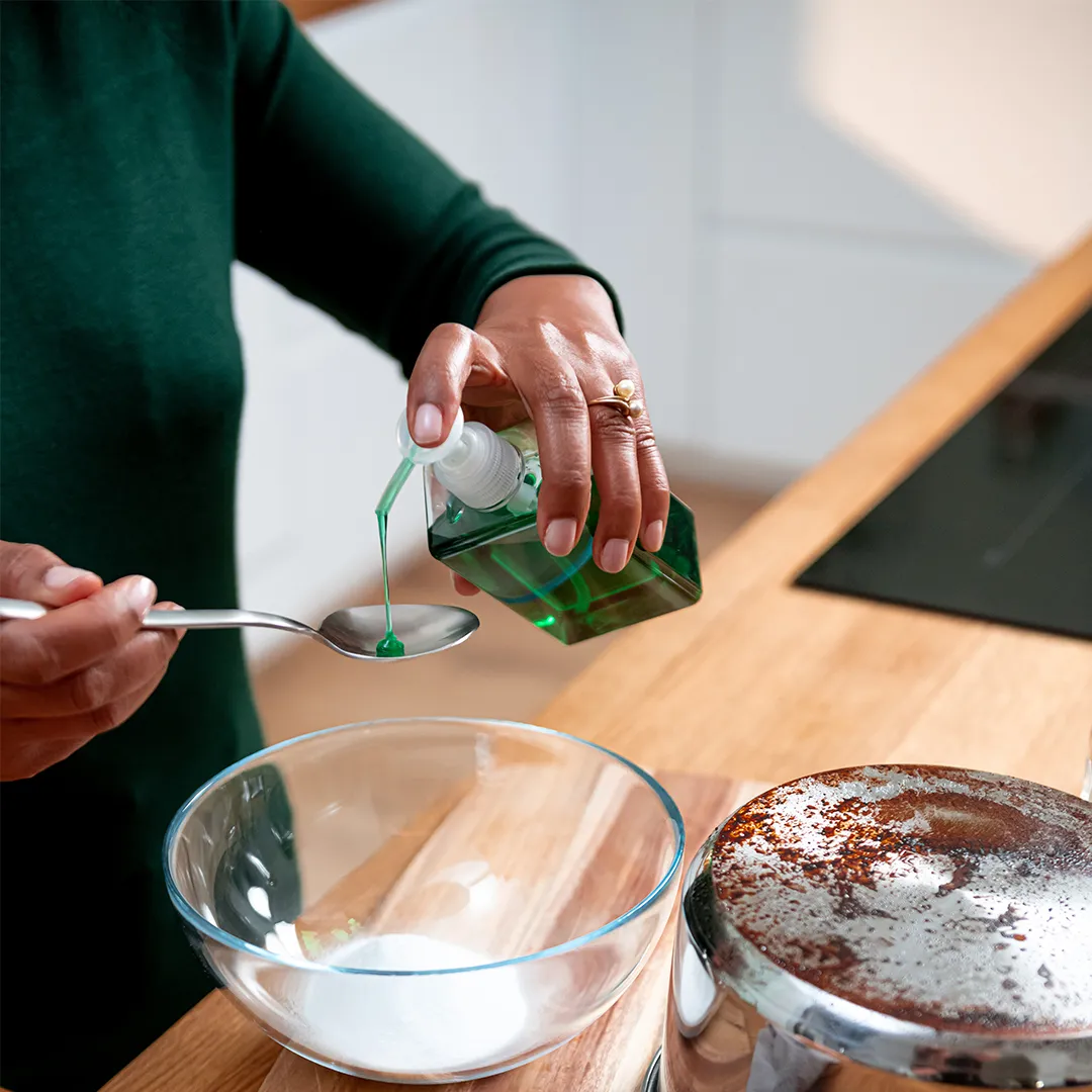 Person pouring green liquid from bottle into spoon over glass bowl in kitchen.