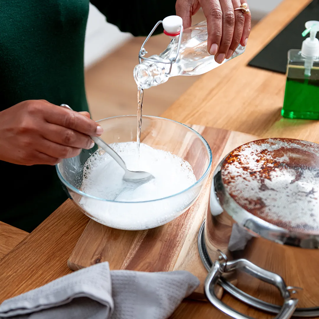 A person pours white vinegar into a glass bowl beside a stainless steel pot on a wooden counter.