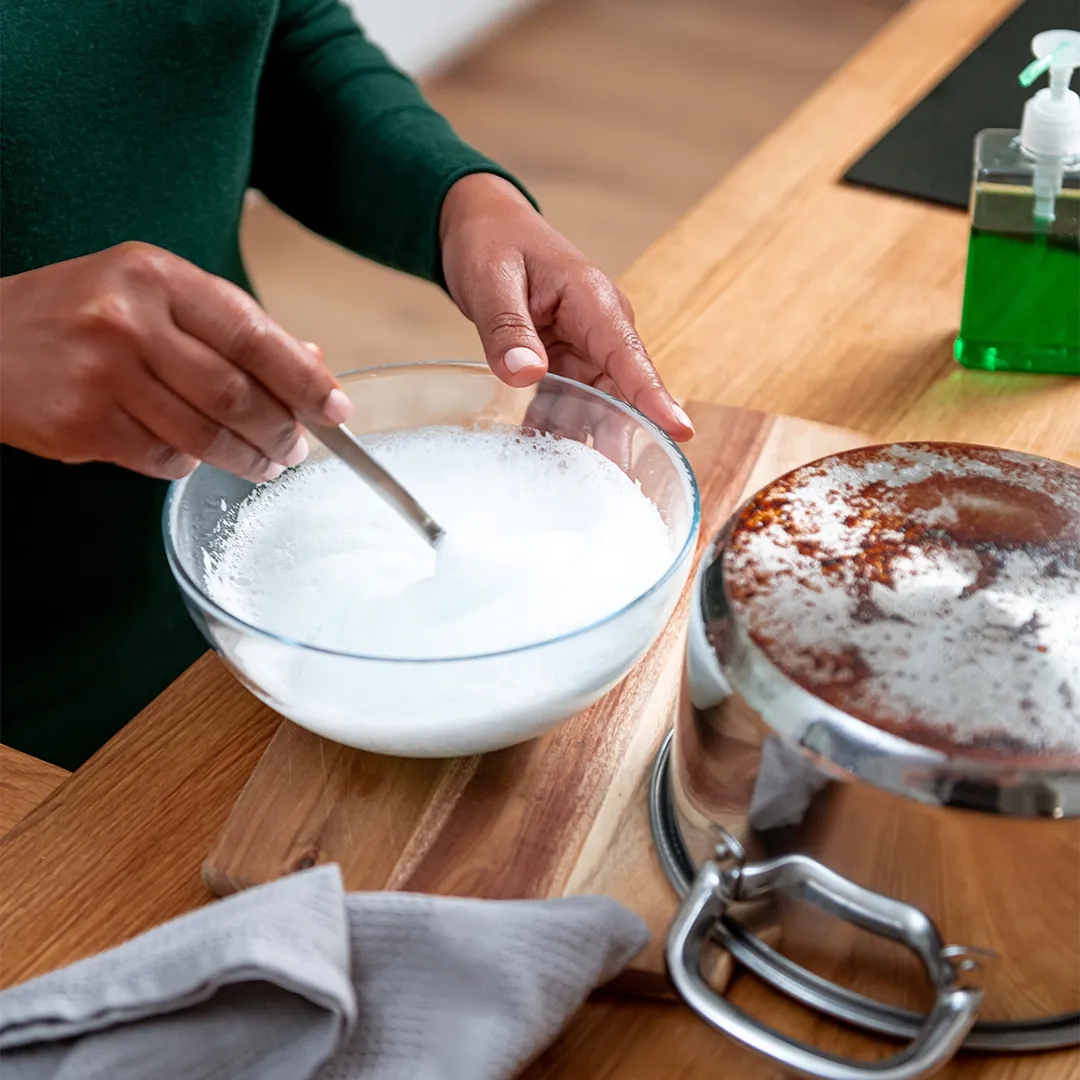 Person stirring white mixture in glass bowl beside a pot with seasoning on a wooden surface.