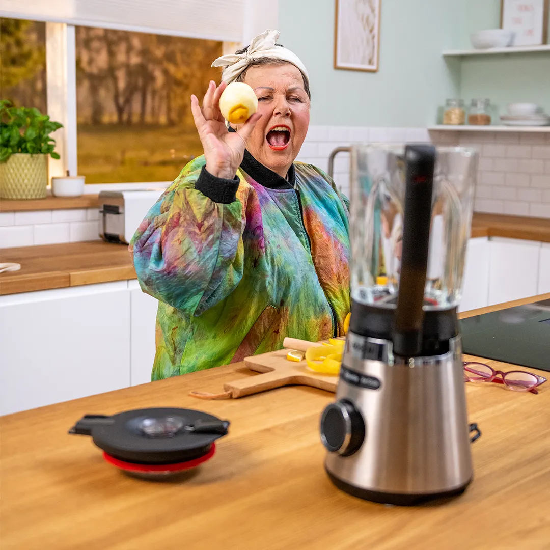 Happy person in colorful shirt holding an apple in a kitchen with a silver blender in front.