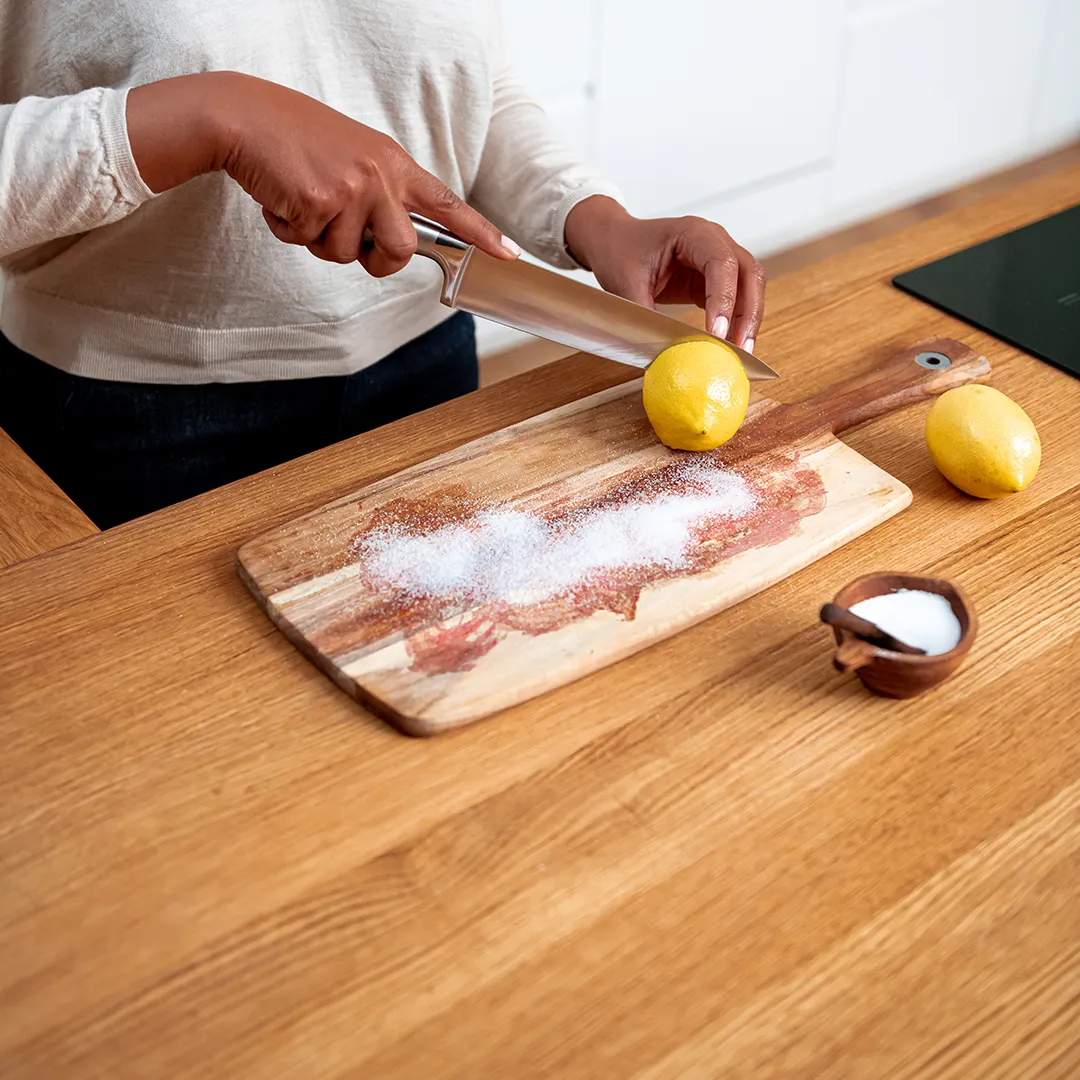 Person slicing a lemon on a wooden cutting board with salt on a kitchen counter.