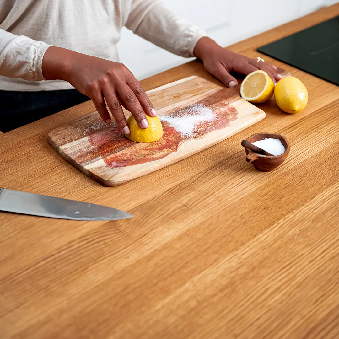 Person cleaning a wooden cutting board with half a lemon and salt on a kitchen counter.