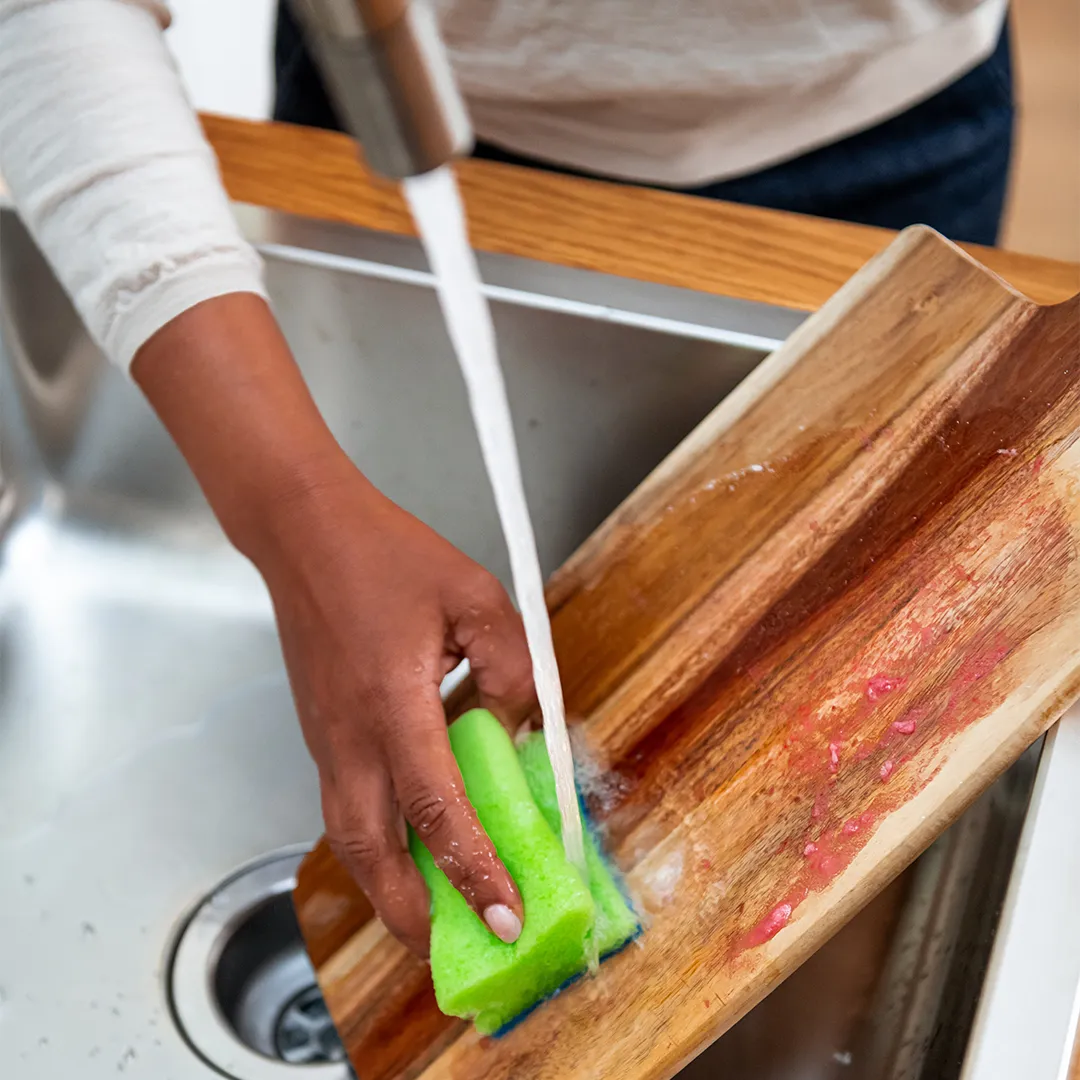 A person cleaning a wooden cutting board with a green sponge under running water in a sink.