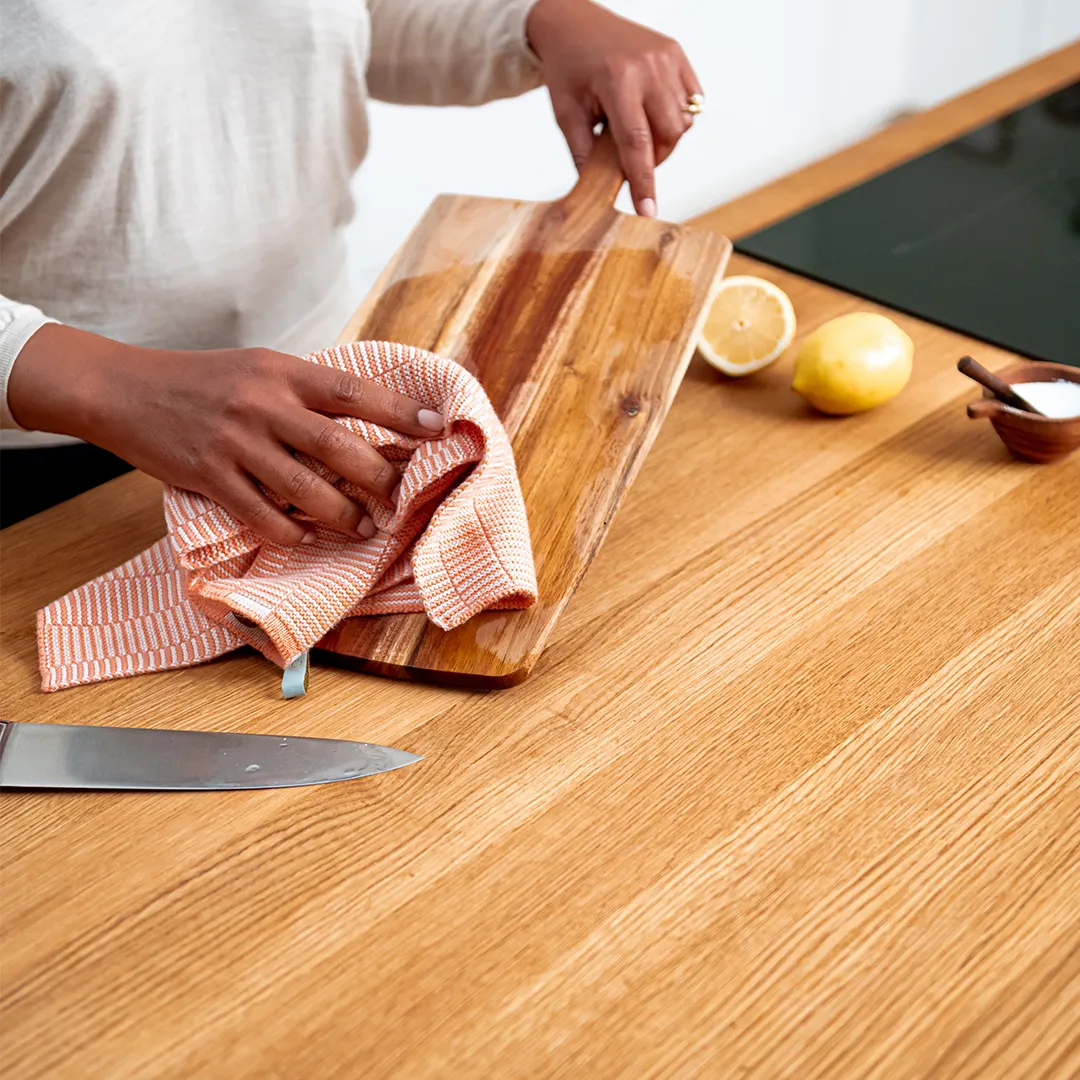 Person cleaning a wooden cutting board with a cloth on a kitchen counter.