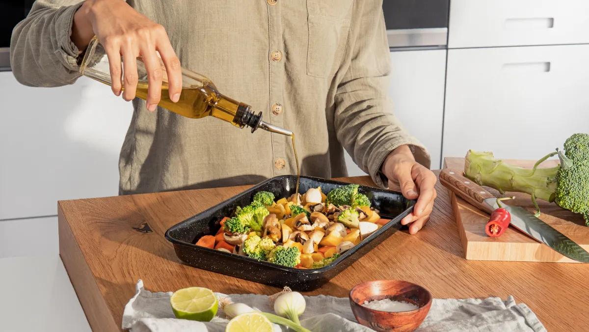 Person drizzling olive oil over a tray of fresh vegetables, preparing a homemade meal in a modern kitchen setting.