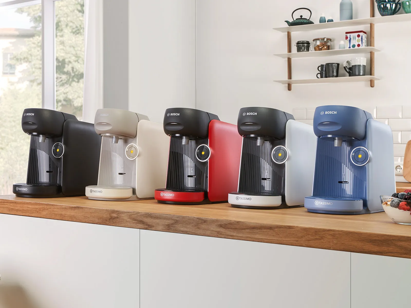 O varietate de cafetiere Bosch Tassimo în negru, bej, roșu și albastru, expuse pe un blat de lemn într-o bucătărie luminoasă.
