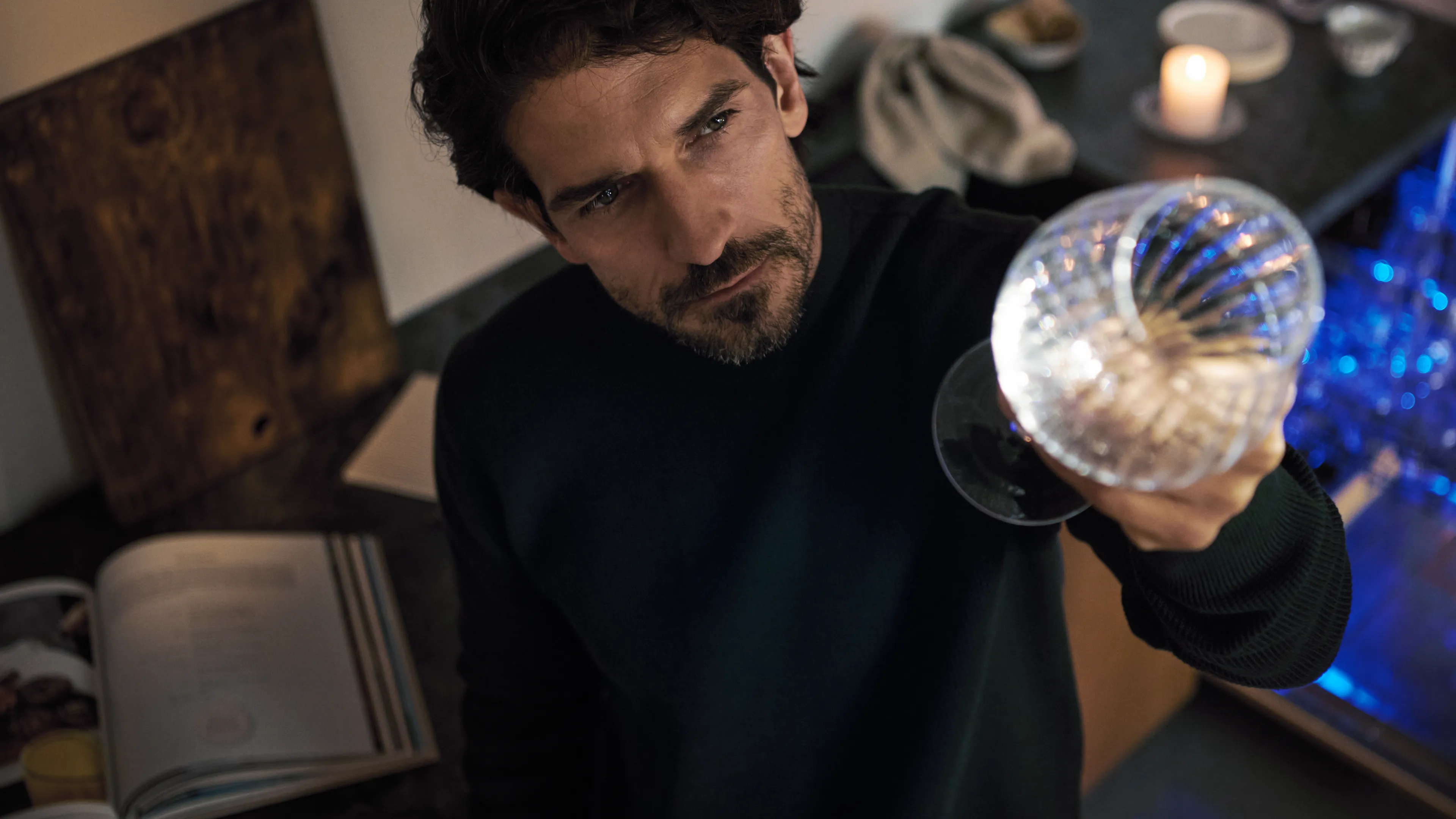 The image shows a man with a beard holding a glass sphere in a dimly lit room, surrounded by books and artwork.