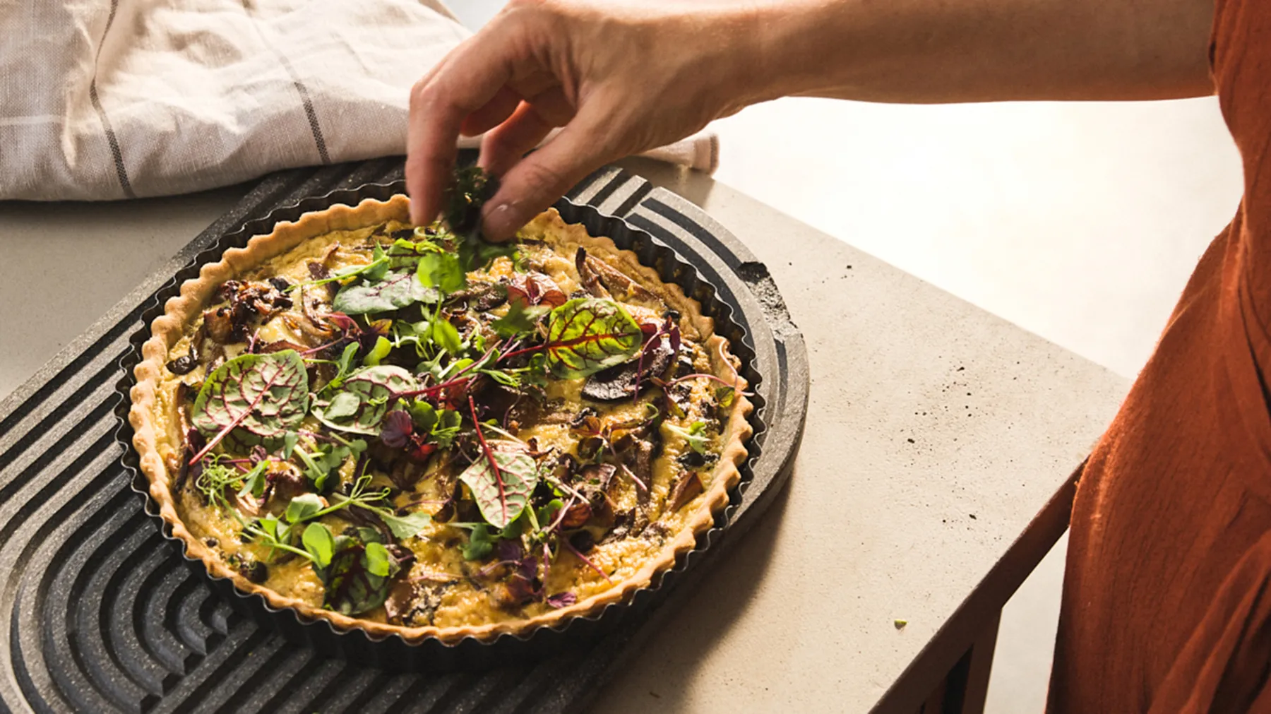 Hands garnishing a savory quiche or tart with fresh greens on a striped platter.