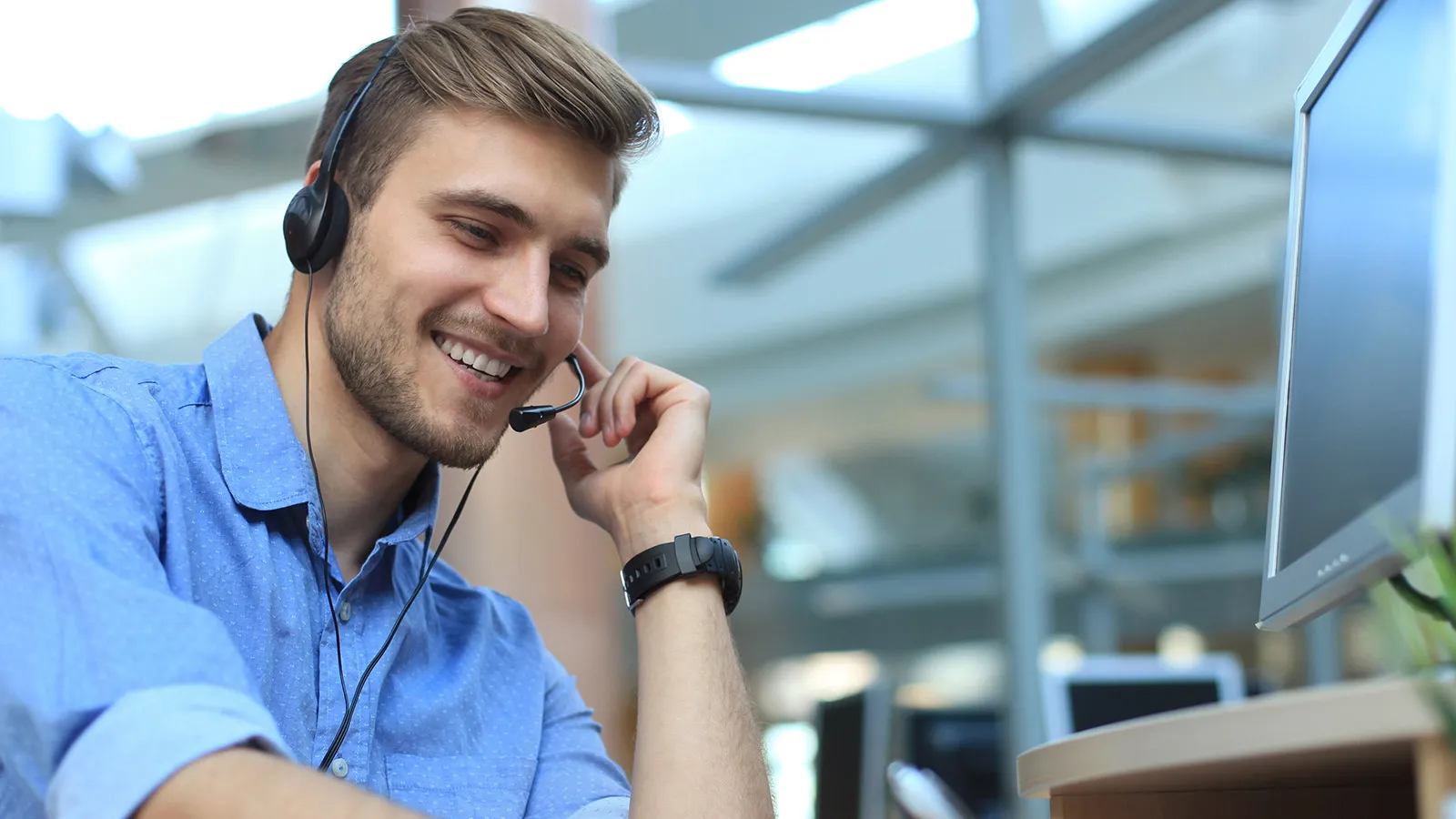 Mlad moški s slušalkami sedi za mizo, osredotočen na računalniški zaslon v svetlem, sodobnem delovnem prostoru.