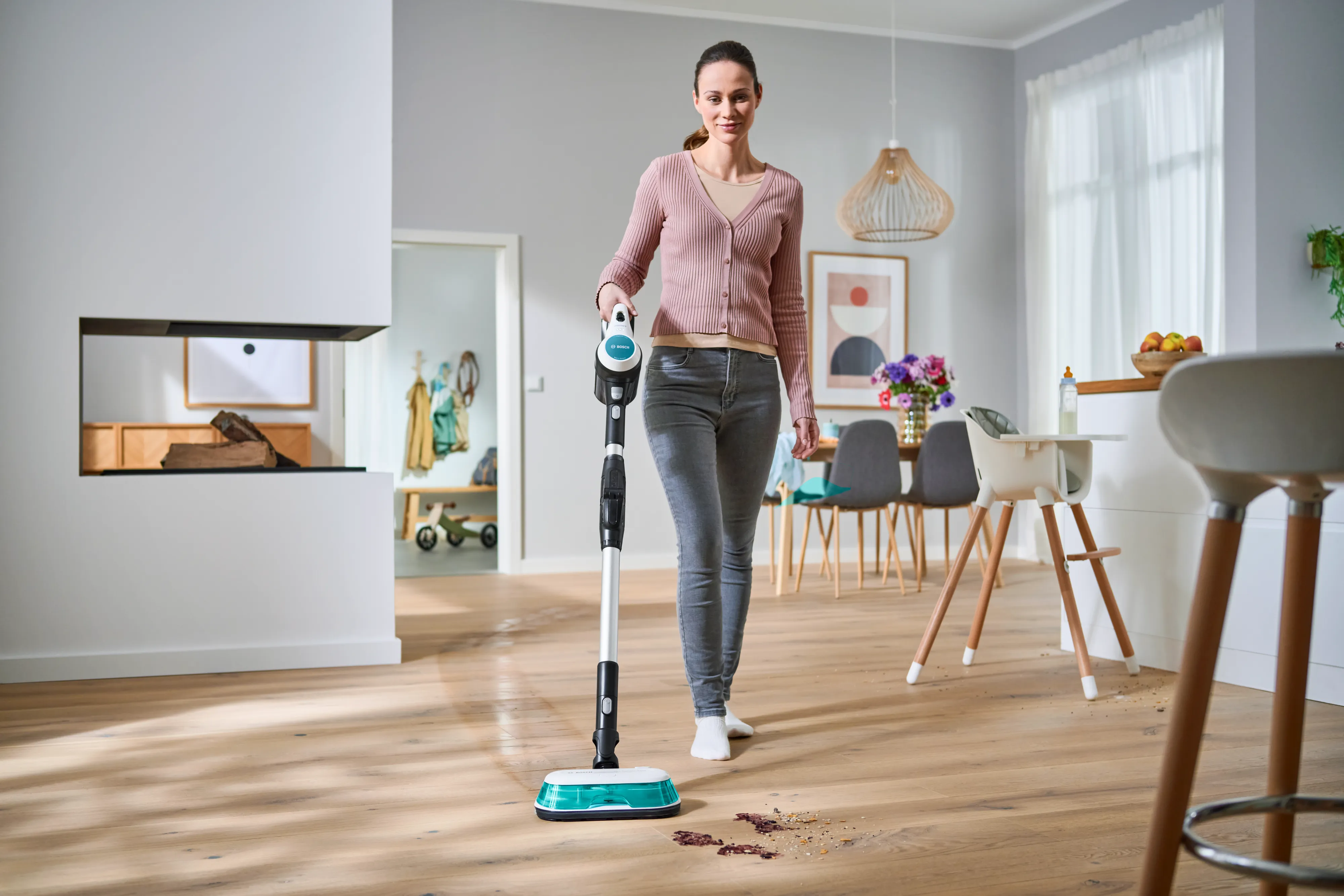 A smiling woman uses a Bosch cordless wet and dry vacuum cleaner to clean up spills and debris on a wooden floor in a bright, modern home.