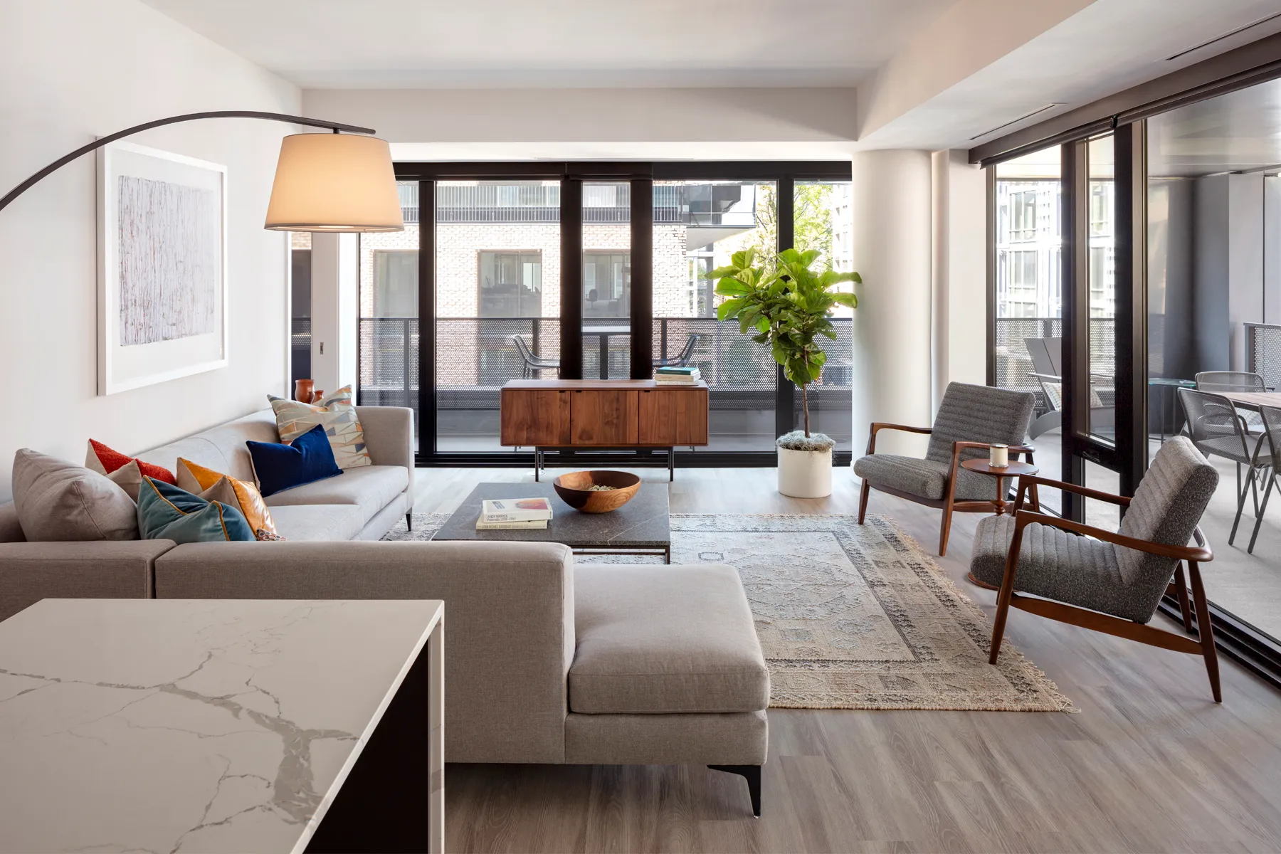 Modern living room with large windows, gray sofa, armchairs, and indoor plant.