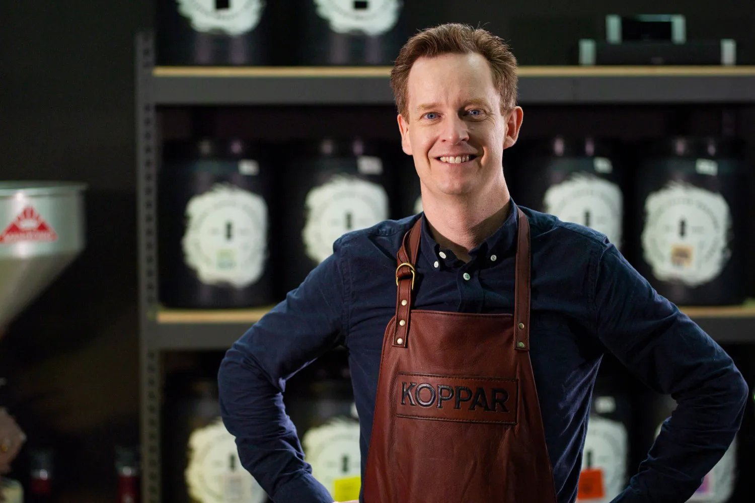 En person i mørk skjorte og forkle står foran hyller med krukker og utstråler profesjonalitet