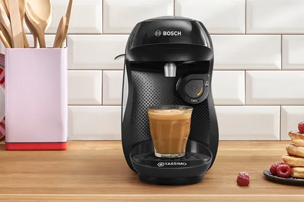 Aparat de cafea TASSIMO cu siglă 57% plastic reciclat, ustensile de bucătărie, și produse coapte pe masă de lemn. 
