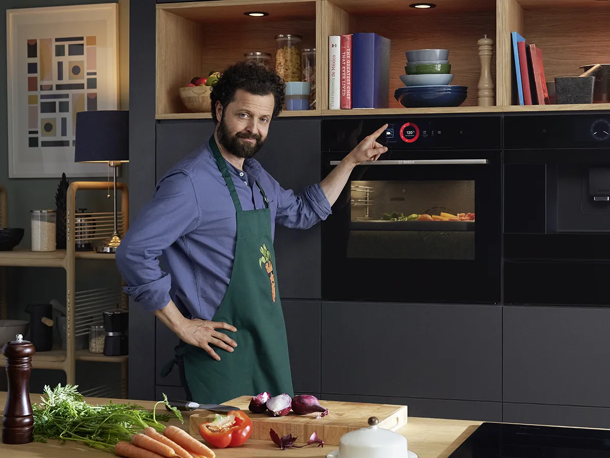 Man in apron standing in kitchen, preparing vegetables on cutting board, oven in background.