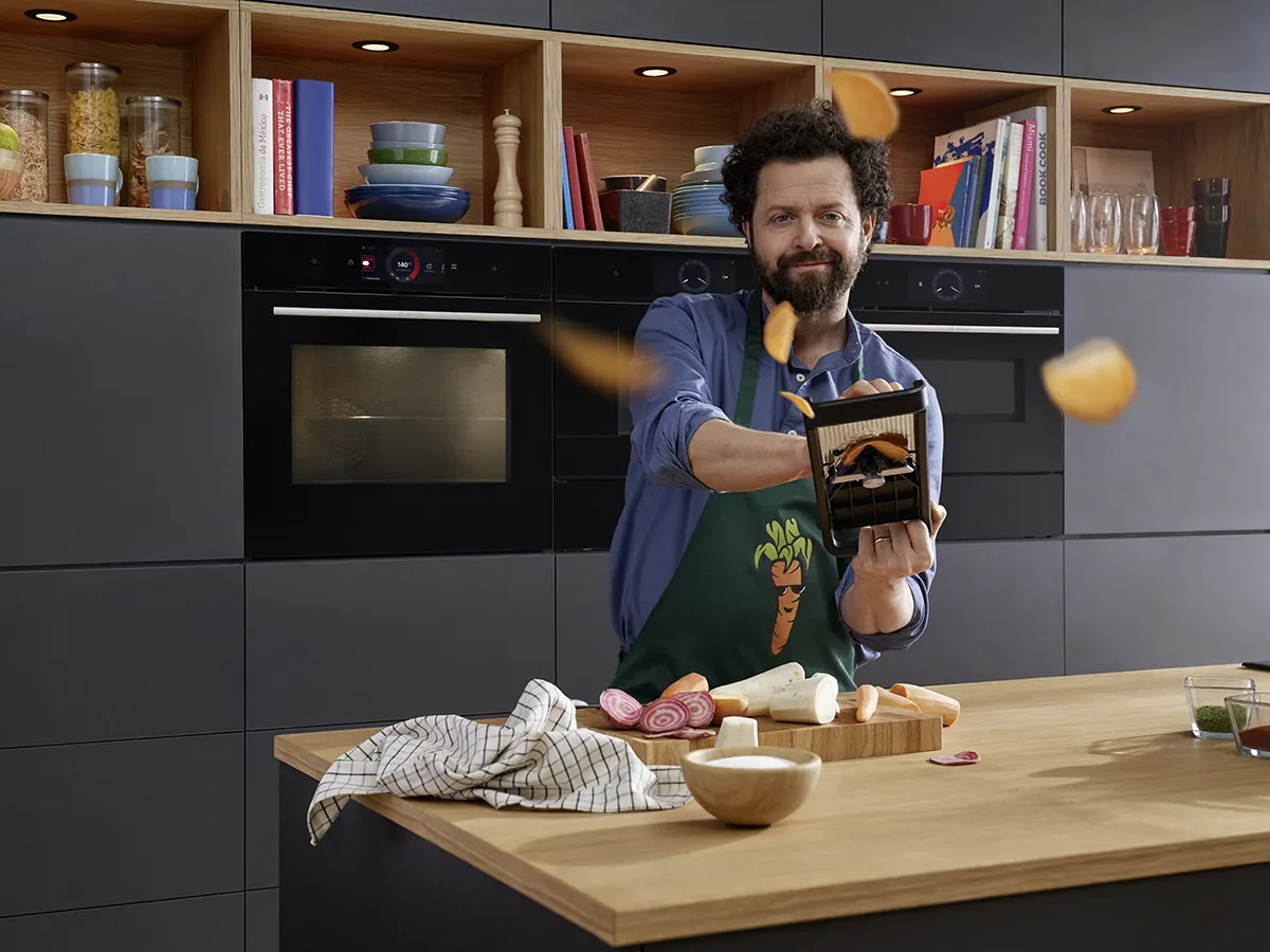 Man in kitchen cooking, holding food item, surrounded by kitchen appliances and utensils.