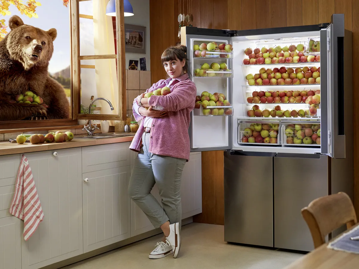 Woman in kitchen with large refrigerator full of apples, standing next to a bear statue.