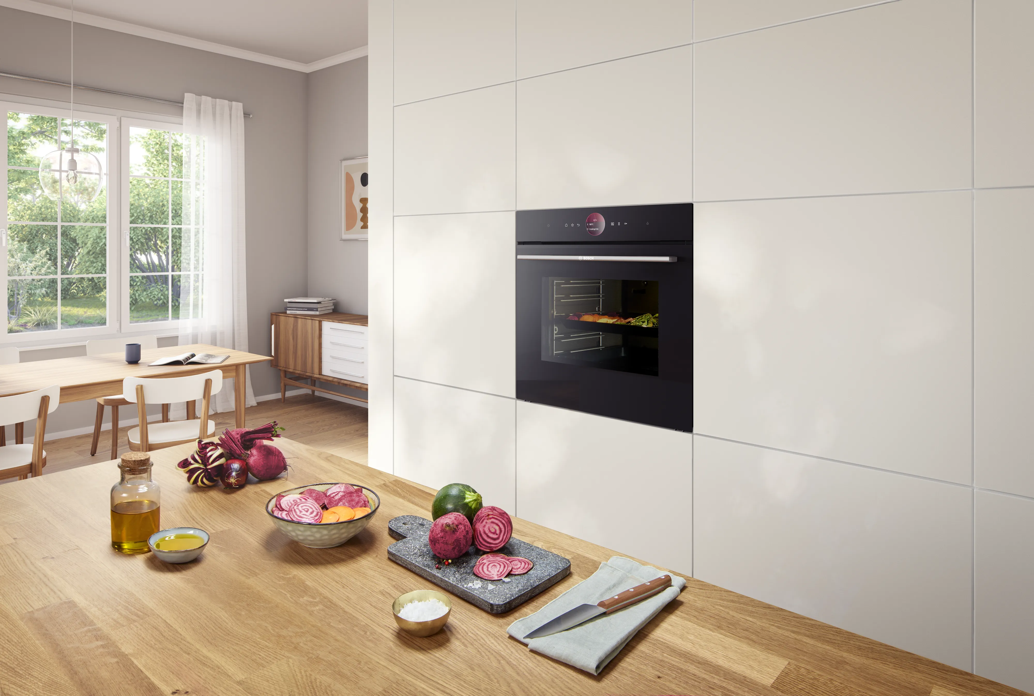 Cucina moderna con forno a incasso, piano di lavoro in legno con barbabietole affettate, olio e utensili da cucina vicino a una finestra con tende bianche trasparenti.