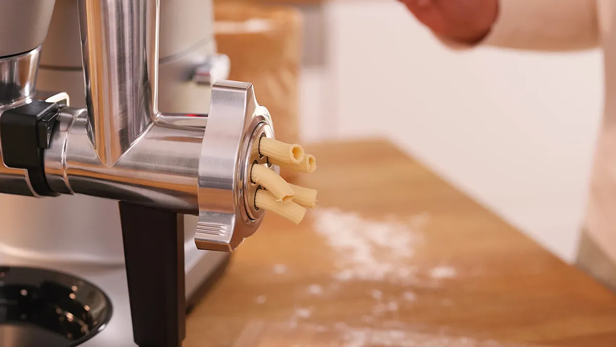 Pasta maker extruding dough, kitchen counter with flour, cooking utensils visible.