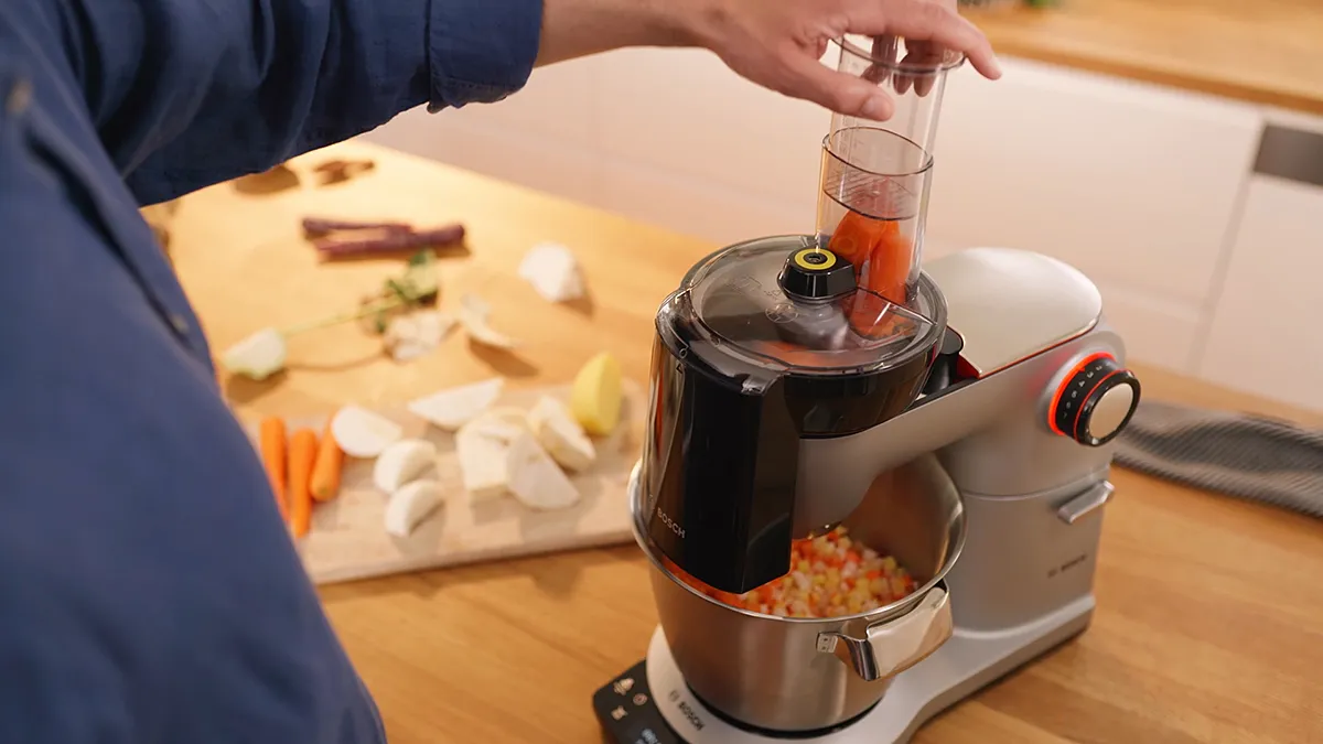 Person using food processor to chop vegetables on wooden table with ingredients.