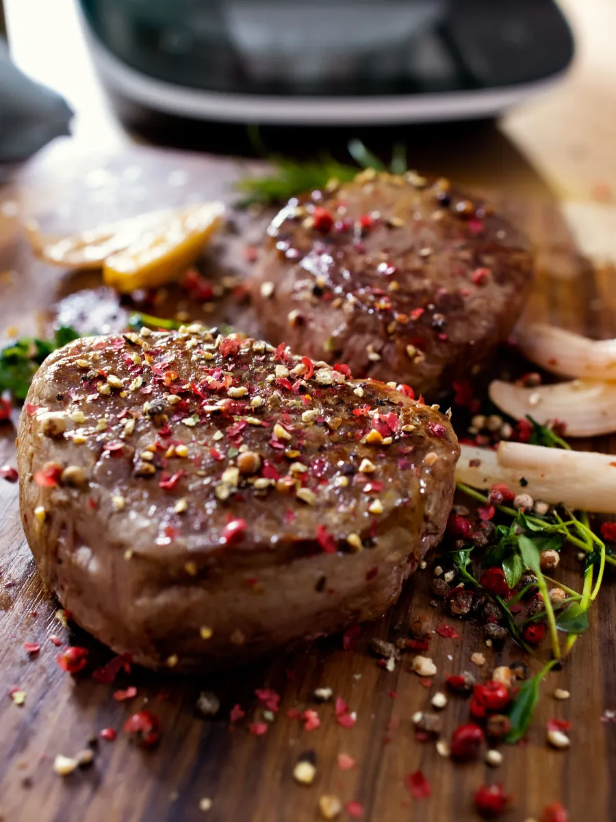 Zwei saftige, perfekt gewürzte Steaks liegen auf einem Holzbrett, umgeben von Kräutern und Gewürzen. Im Hintergrund ist die Bosch Cookit Küchenmaschine zu sehen.