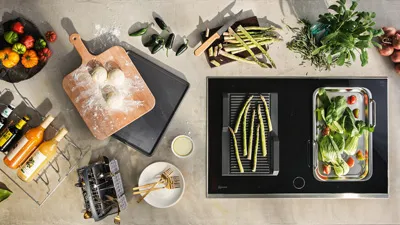alt-A modern kitchen setup with NEFF cooking accessories, fresh vegetables, and colorful drinks arranged around a sleek cooking surface.