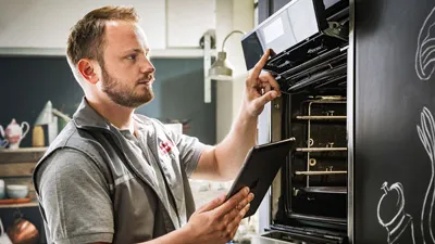 alt-A NEFF technician wearing gloves works on a modern oven, using a screwdriver against a backdrop of home decor and green walls.