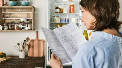 alt-A NEFF customer stands in a stylish kitchen, reading a user manual, surrounded by colorful ingredients and kitchenware.