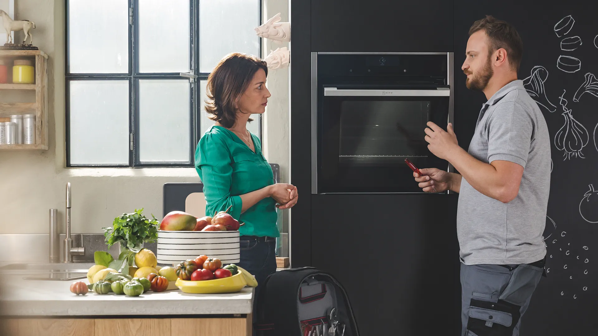 A NEFF technician in a gray polo shirt stands in a modern kitchen, interacting with a tablet near a sleek cooktop and bowls of fresh fruit.
