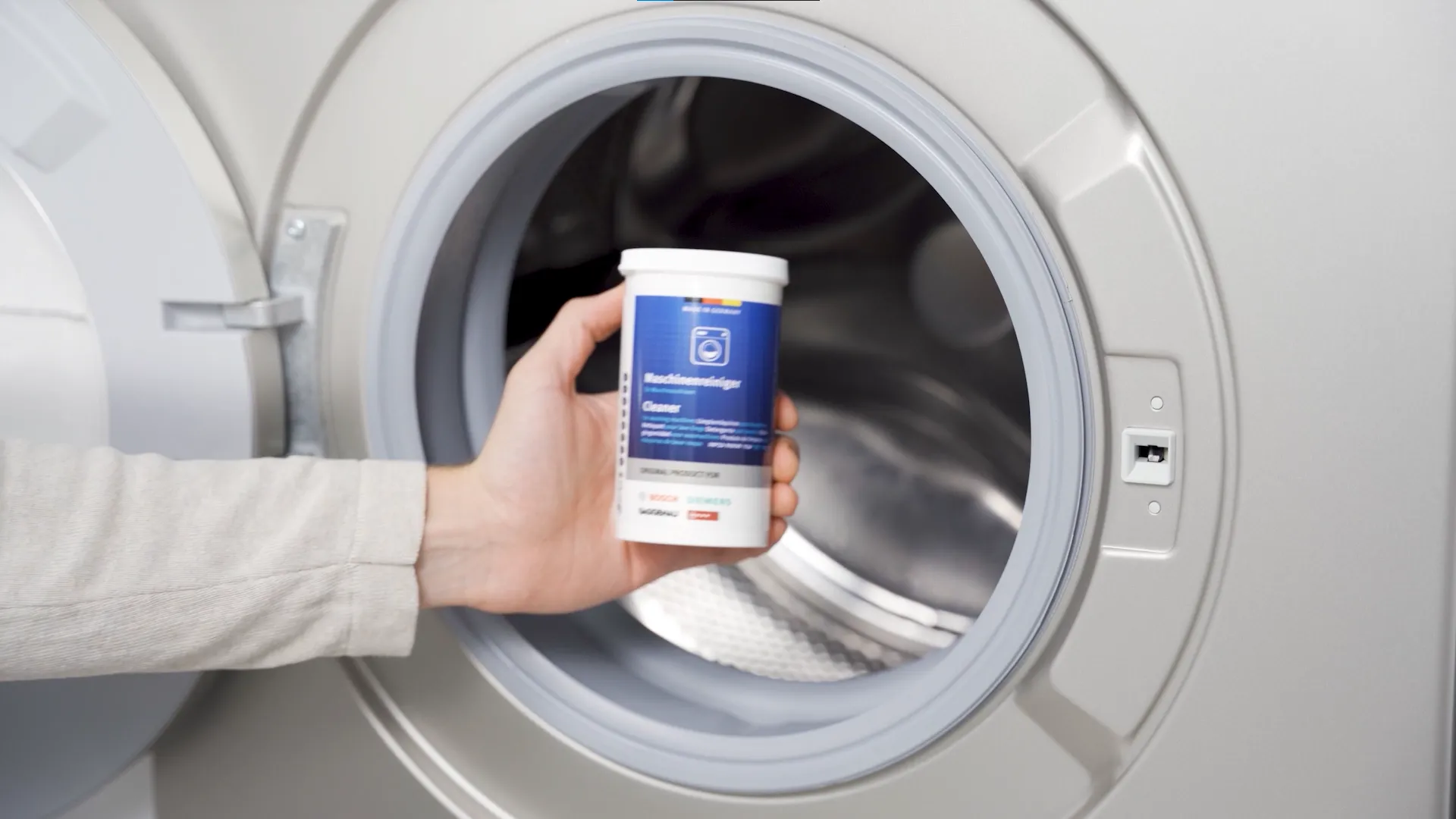 Hand holding Bosch washing machine cleaner in front of open washing machine drum.