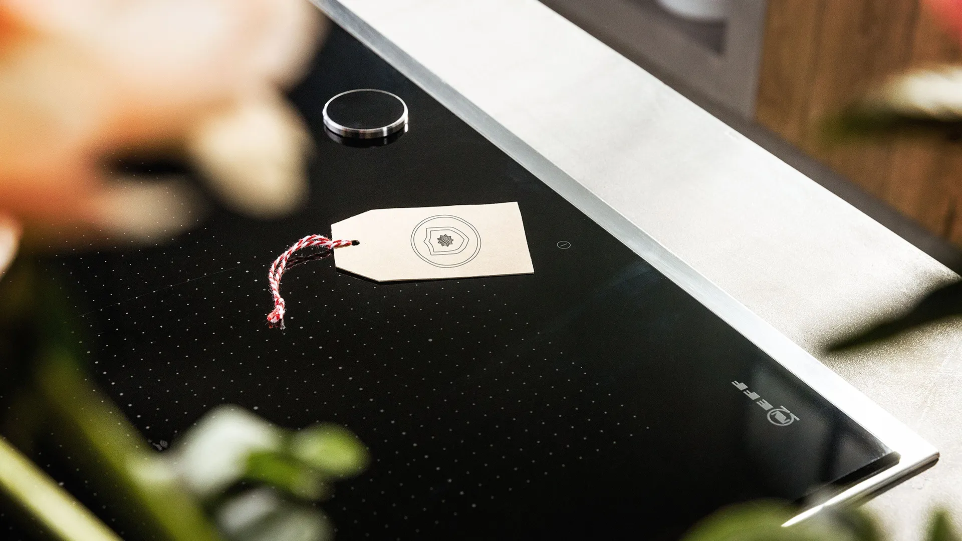 Een beige label met een rood-wit koordje op een zwarte NEFF kookplaat, met onscherpe planten in de voorgrond.