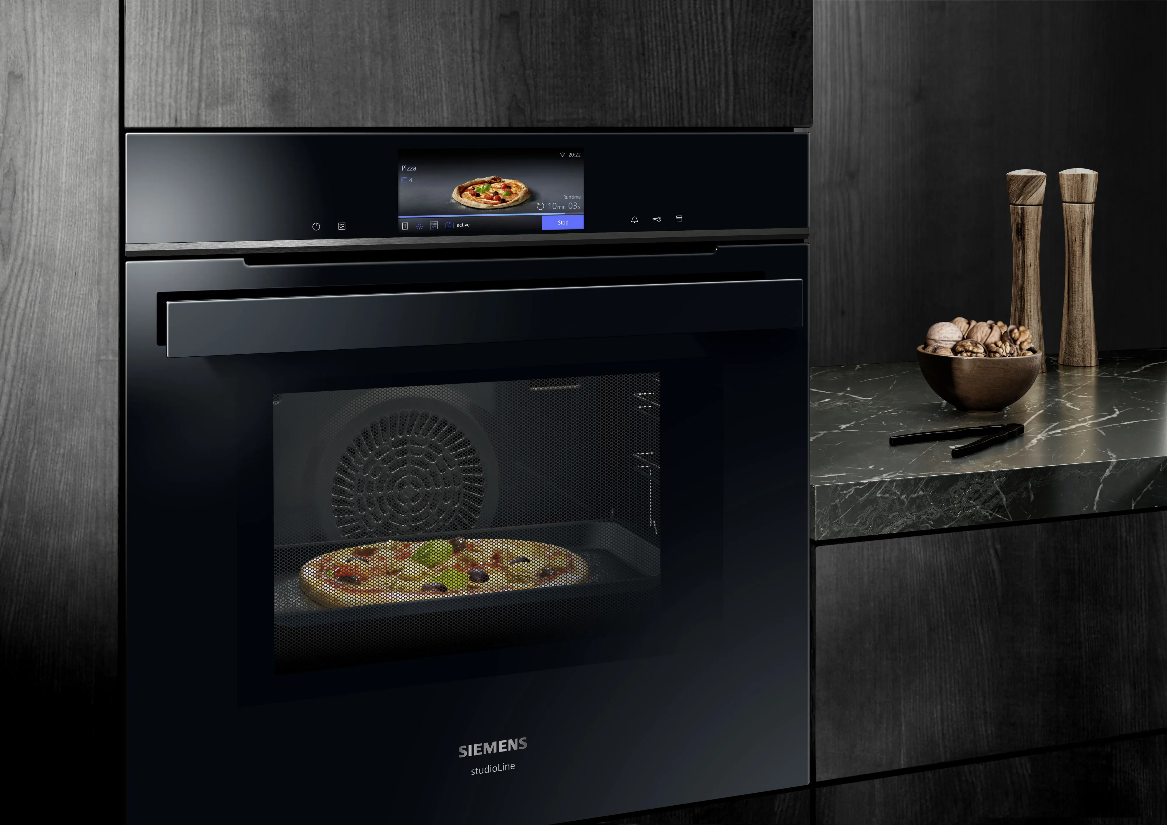 Forno embutido preto com pizza dentro, bancada de mármore, utensílios de cozinha, armários de madeira escura.